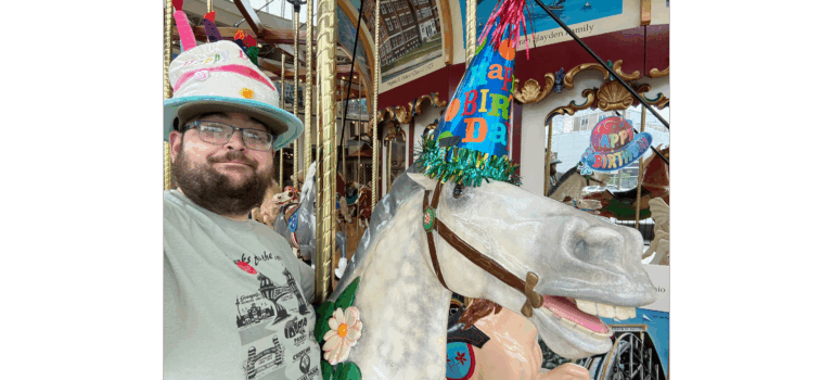 Kevin on the Euclid Beach Grand Carousel at the Cleveland History Center. Both Kevin and the pictured carousel horse are wearing party hats.